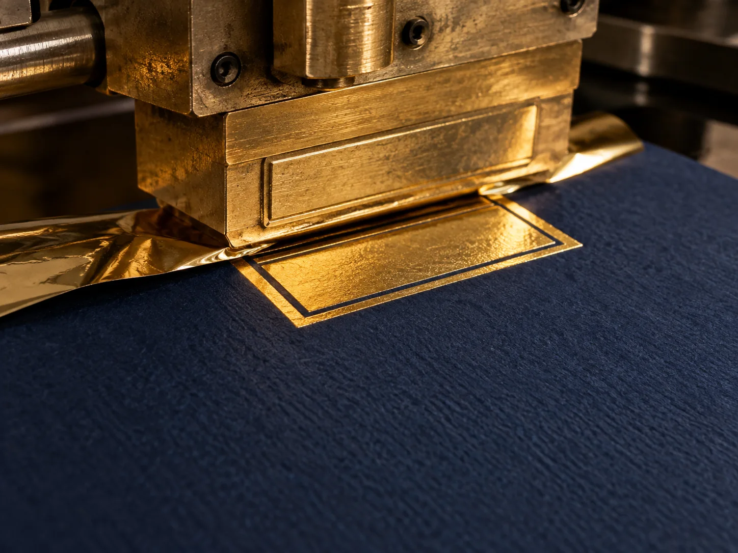 Heated brass die pressing reflective gold foil onto textured navy paper